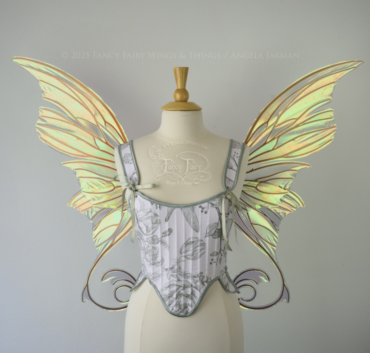 Large light green & orange iridescent fairy wings with lots of intricate veins, some with 'thorns'. The upper panels curve slightly downwards along the top edge with pointy tips, the bottom panels have tails that curve inward toward the body. Worn on a dress form which is also wearing white & sage green floral stays / bodice