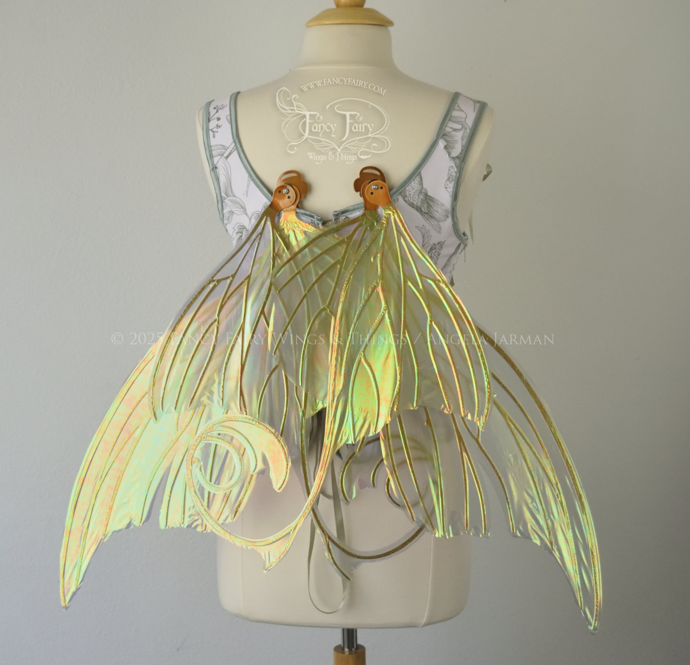 Large light green & orange iridescent fairy wings with lots of intricate veins, some with 'thorns'. The upper panels curve slightly downwards along the top edge with pointy tips, the bottom panels have tails that curve inward toward the body. Worn on a dress form which is also wearing white & sage green floral stays / bodice, back view, in resting position