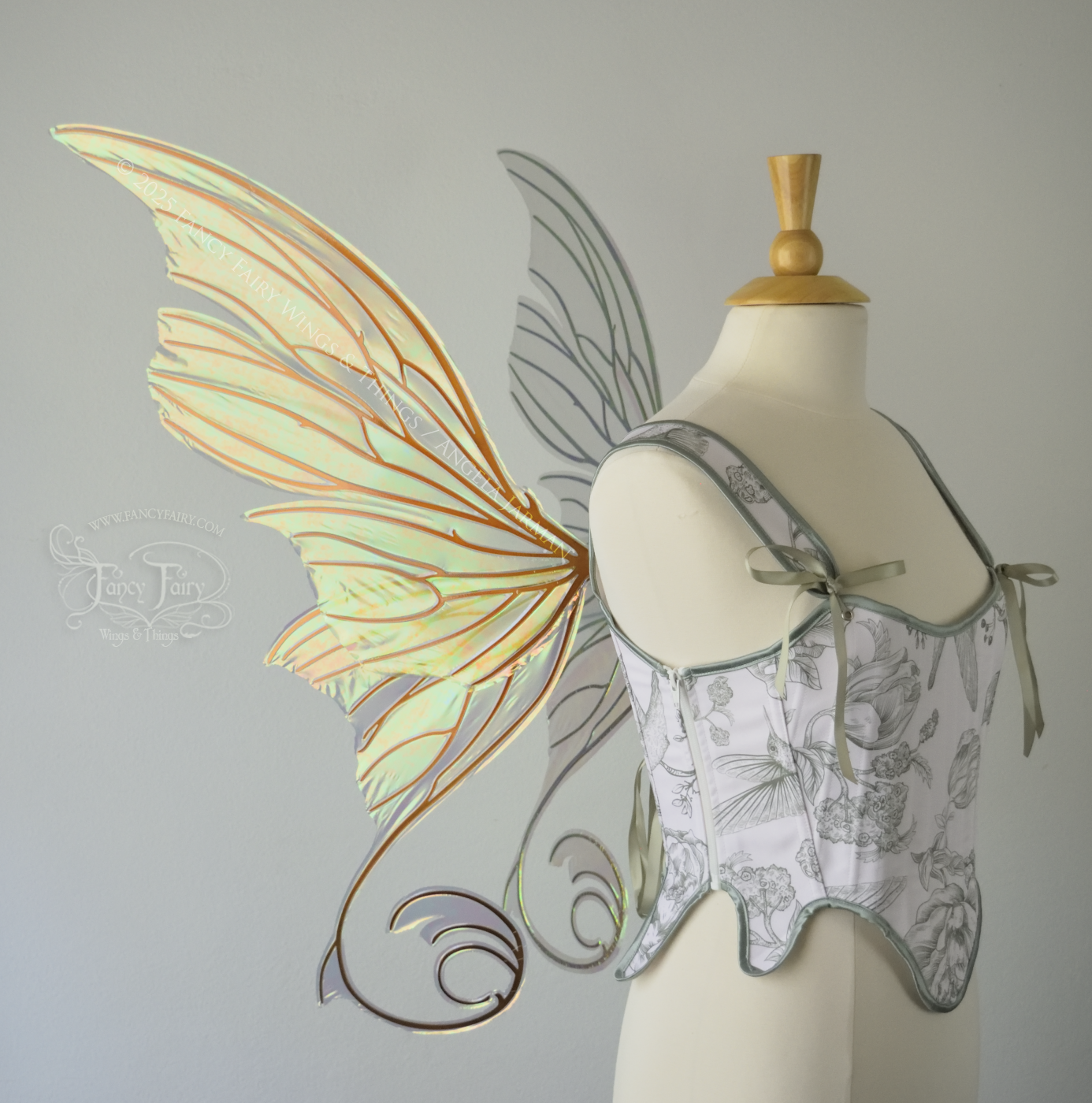 Large light green & orange iridescent fairy wings with lots of intricate veins, some with 'thorns'. The upper panels curve slightly downwards along the top edge with pointy tips, the bottom panels have tails that curve inward toward the body. Worn on a dress form which is also wearing white & sage green floral stays / bodice, left side view