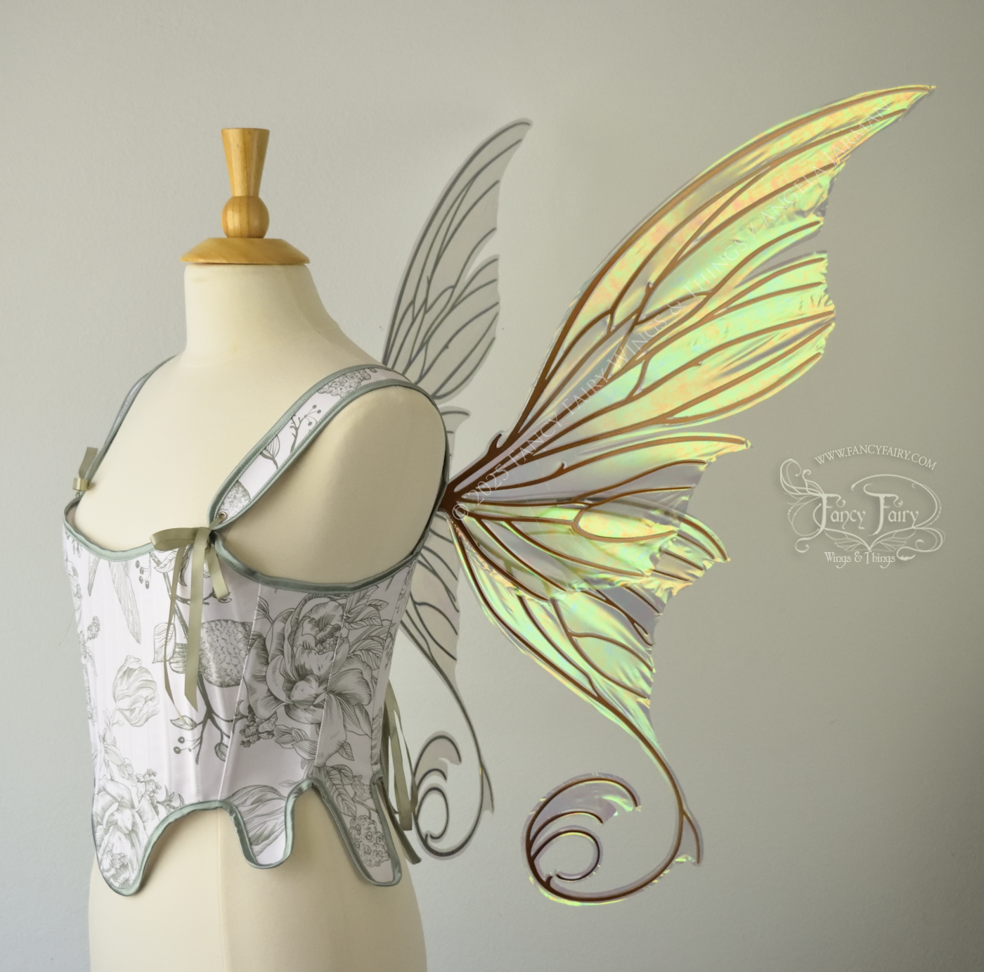 Large light green & orange iridescent fairy wings with lots of intricate veins, some with 'thorns'. The upper panels curve slightly downwards along the top edge with pointy tips, the bottom panels have tails that curve inward toward the body. Worn on a dress form which is also wearing white & sage green floral stays / bodice, right side view