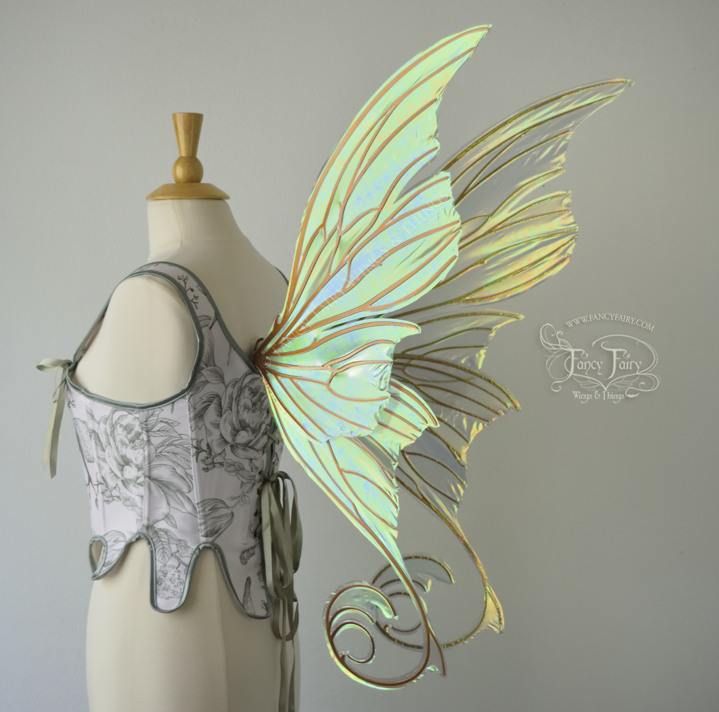 Large light green & orange iridescent fairy wings with lots of intricate veins, some with 'thorns'. The upper panels curve slightly downwards along the top edge with pointy tips, the bottom panels have tails that curve inward toward the body. Worn on a dress form which is also wearing white & sage green floral stays / bodice, back 3/4 view