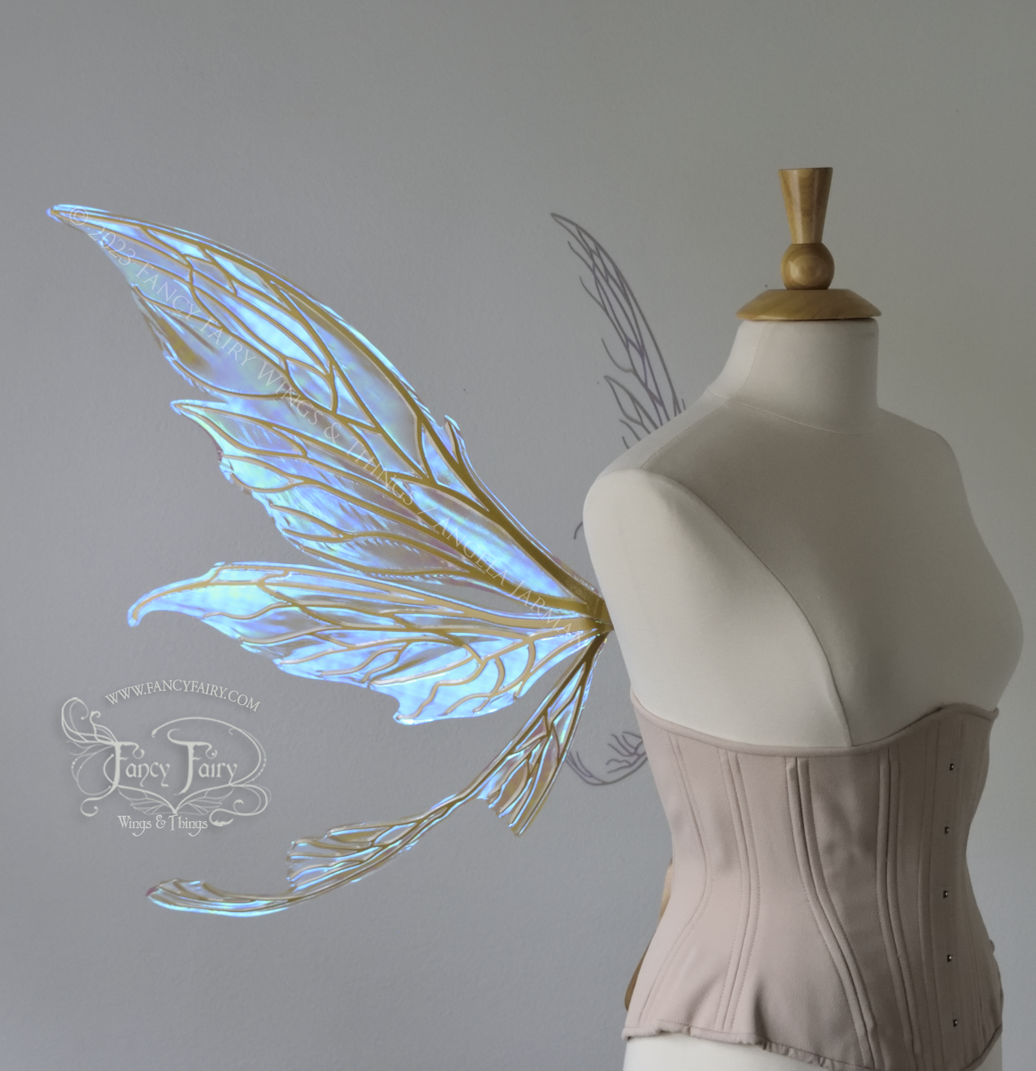 Right side view of a dress form wearing an underbust corset & large blue iridescent fairy wings with gold veins. Upper panels are elongated with pointed tips, curved ‘tail’