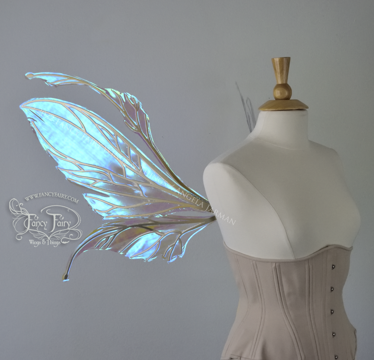 Left side view of an ivory dress form wearing an alabaster underbust corset and large fairy wings featuring antennae along the top. The upper panels are elongated with semi-pointed tips, the lower panels are smaller with slender tips that curve downward. They are transparent iridescent with a mostly blue shine with some green and the veins are metallic gold. The background is plain white and my logo and copyright notice are visible.