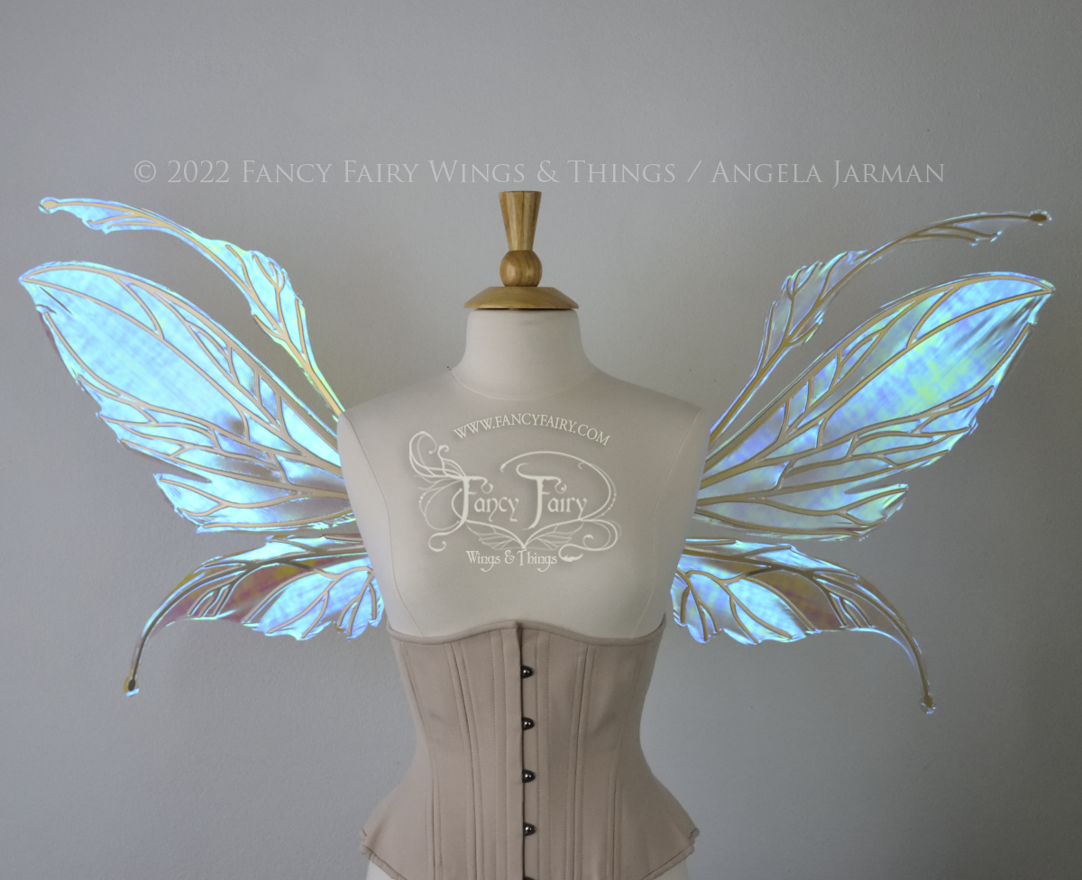 Front view of an ivory dress form wearing an alabaster underbust corset and large fairy wings featuring antennae along the top. The upper panels are elongated with semi-pointed tips, the lower panels are smaller with slender tips that curve downward. They are transparent iridescent with a mostly blue shine with some green and the veins are metallic gold. The background is plain white and my logo and copyright notice are visible.