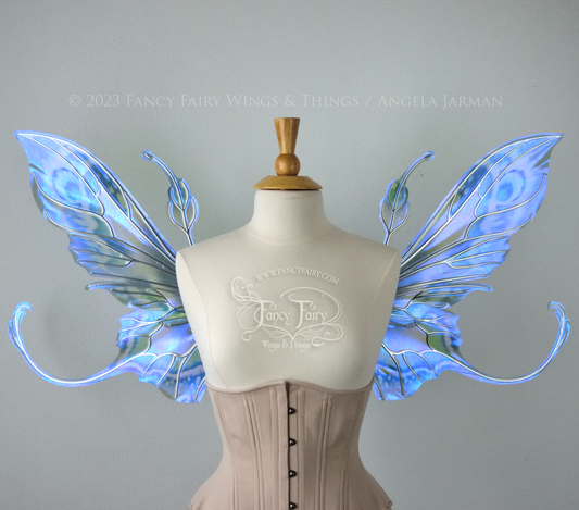 Front view of large blue, violet & teal painted iridescent fairy wings, elongated upper panels with antennae, bottom panels have a tail curving upwards, silver veins, worn on a dress form