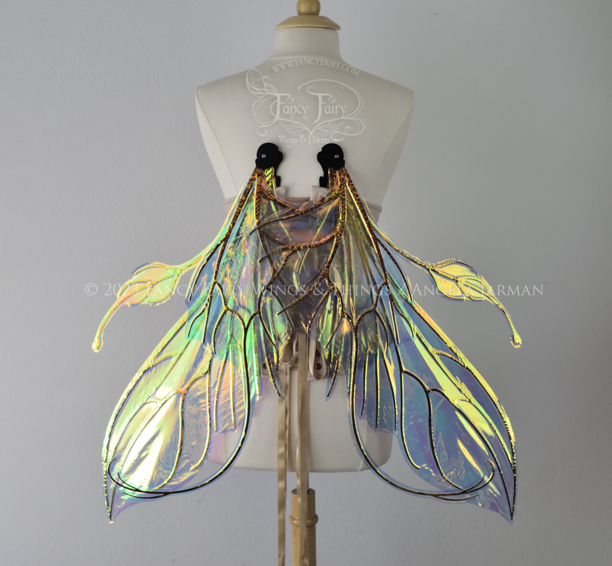 Back view of an ivory dress form wearing an alabaster underbust corset & extra large iridescent fairy wings (in resting position pointed downward) with elongated upper panels & antennae with bottom panels that have a tail curving upwards, black veins. Background is white / light grey