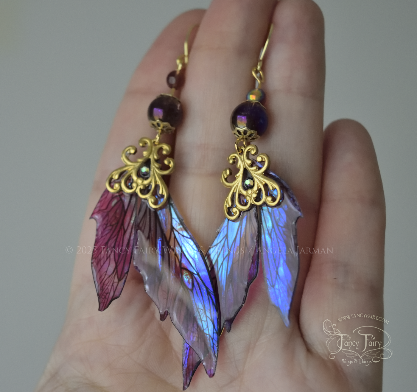 Iridescent fairy wing earrings in purple with brass filigree are held in the palm of a white woman's hand