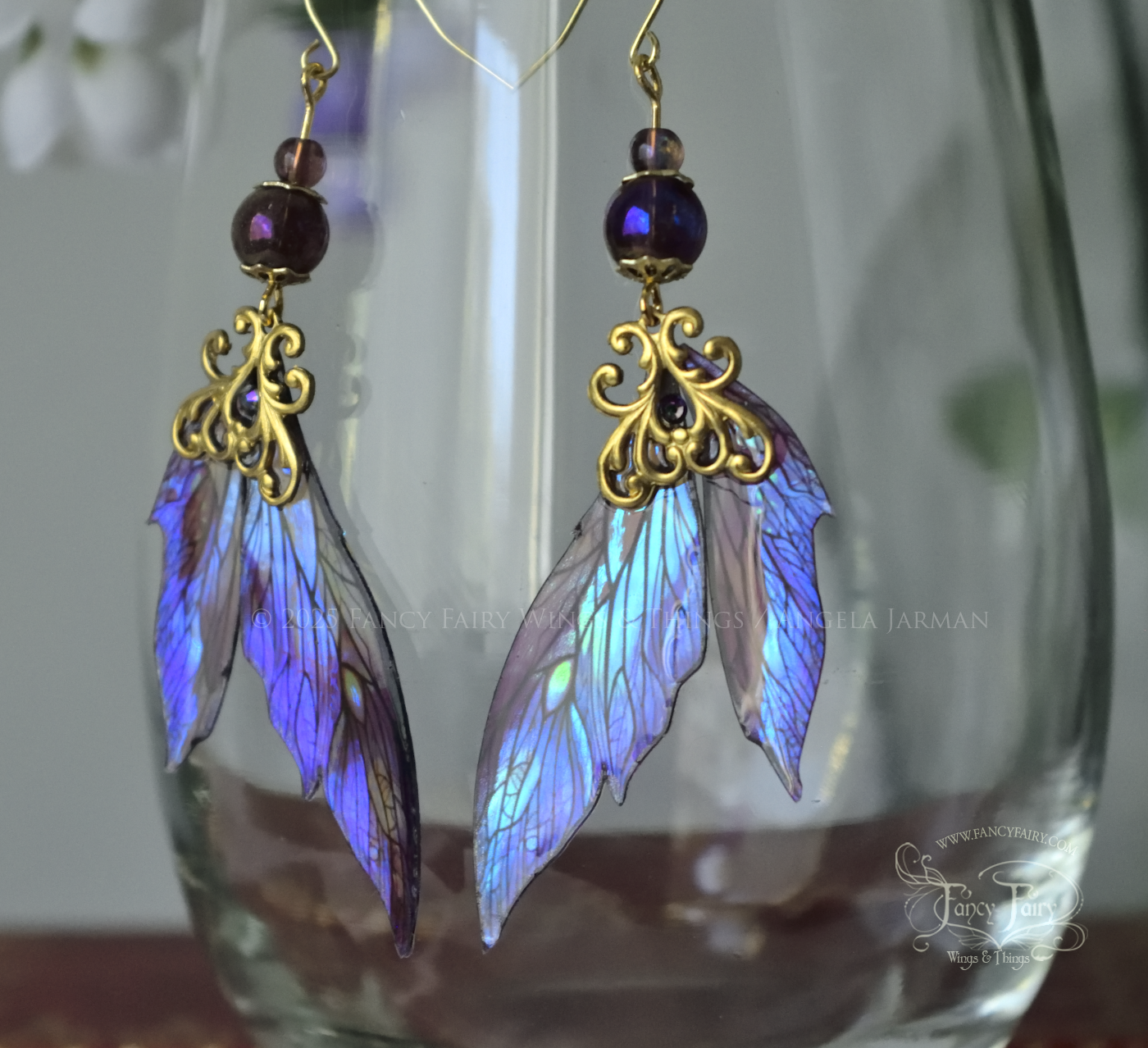 Iridescent fairy wing earrings in purple with brass filigree dangle from the rim of a clear glass