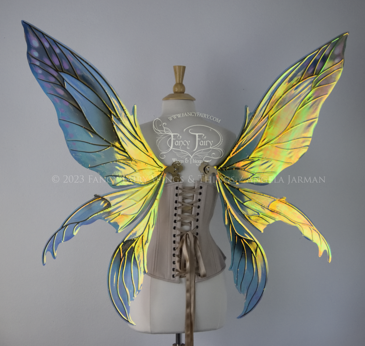 Back view of large fairy wings in iridescent green and blue with pink spots near the tips of the upper panels, with gold veins, worn on a dressform