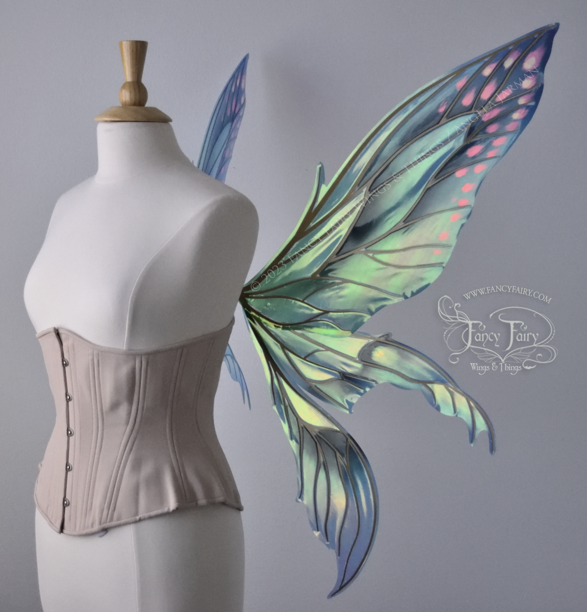 Right side view of large fairy wings in iridescent green and blue with pink spots near the tips of the upper panels, with gold veins, worn on a dressform