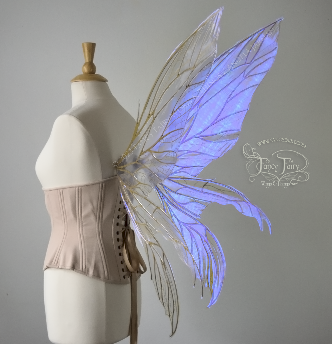 Back 3/4 view of an ivory dress form wearing an alabaster underbust corset & large violet iridescent fairy wings. Upper panels are elongated with pointed tips, 2 lower panels curve downward, lots of thin vein detail in gold