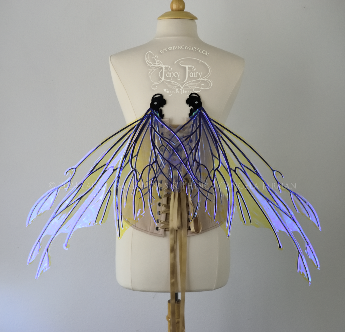 Back view of an ivory dress form wearing an alabaster underbust corset & large violet iridescent fairy wings with elongated upper panels with petal like appendages at the tips & antennae, the veins are black, in resting position