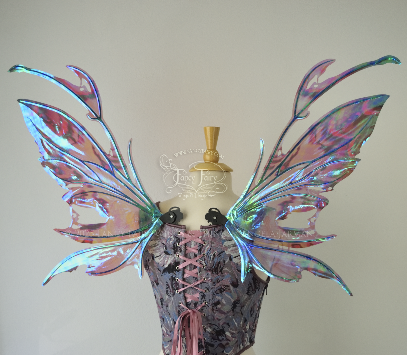 Iridescent fairy wings in mauve, violet, red & black with spikey antennae & tattered edges with holes is worn on a dress form which is also wearing violet & pink floral patterned stays / bodice, back view