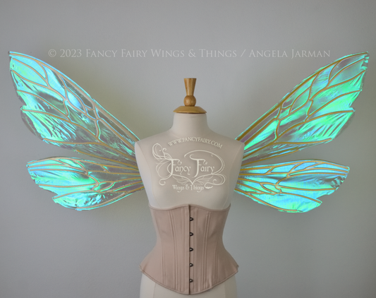 Front view of an ivory dress form wearing an alabaster underbust corset and extra large fairy wings similar to wasp wings, upper and lower panels are both elongated with rounded and slightly pointed tips. They are iridescent green / blue, with gold veins. The background is plain white and my logo and copyright notice are visible.