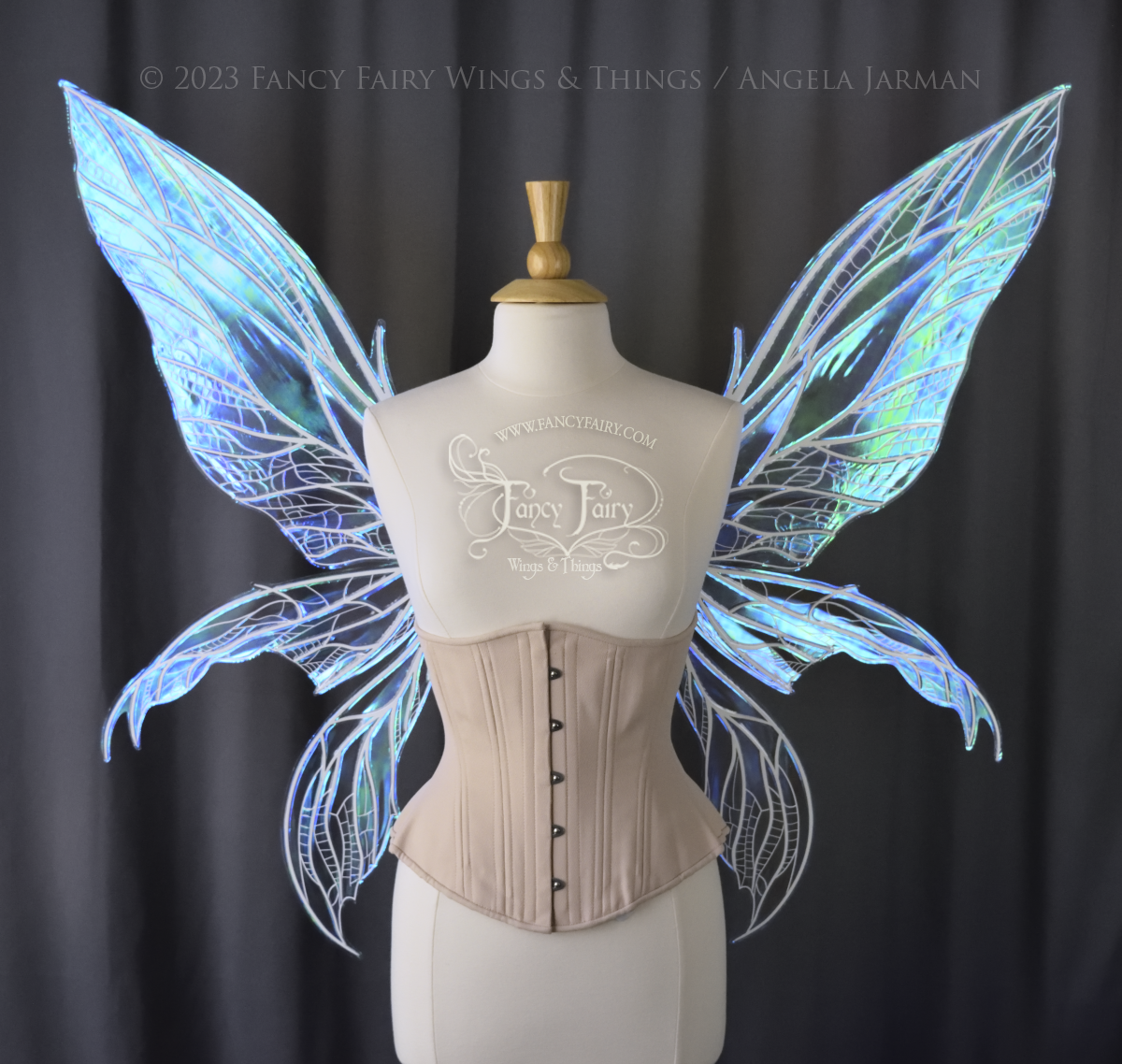 Front view of an ivory dress form wearing an underbust corset and large blue/teal/violet iridescent fairy wings. Upper panels are elongated with pointed tips, 2 lower panels curve downward, detailed white veins, grey background