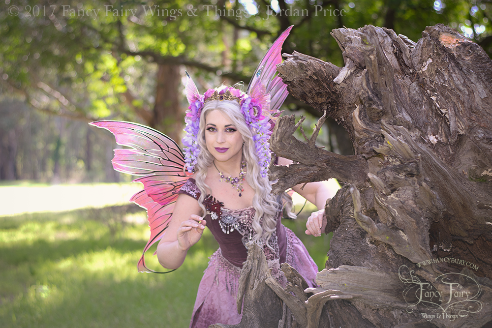 Photo of Angela Jarman, a white woman with long wavy white hair dressed as a fairy with a purple & pink costume & wings is peeking out from behind the roots of a uprooted tree trunk