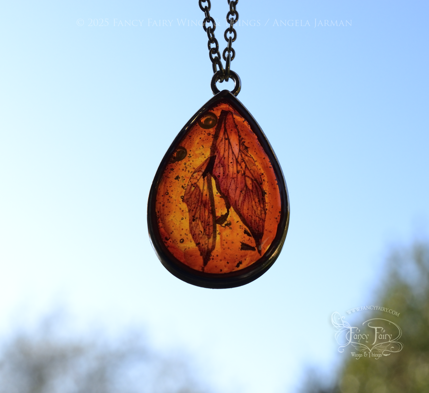 Closeup of faux amber pendant with iridescent fairy wings trapped inside, hanging in front & back lit by a bright light blue sky