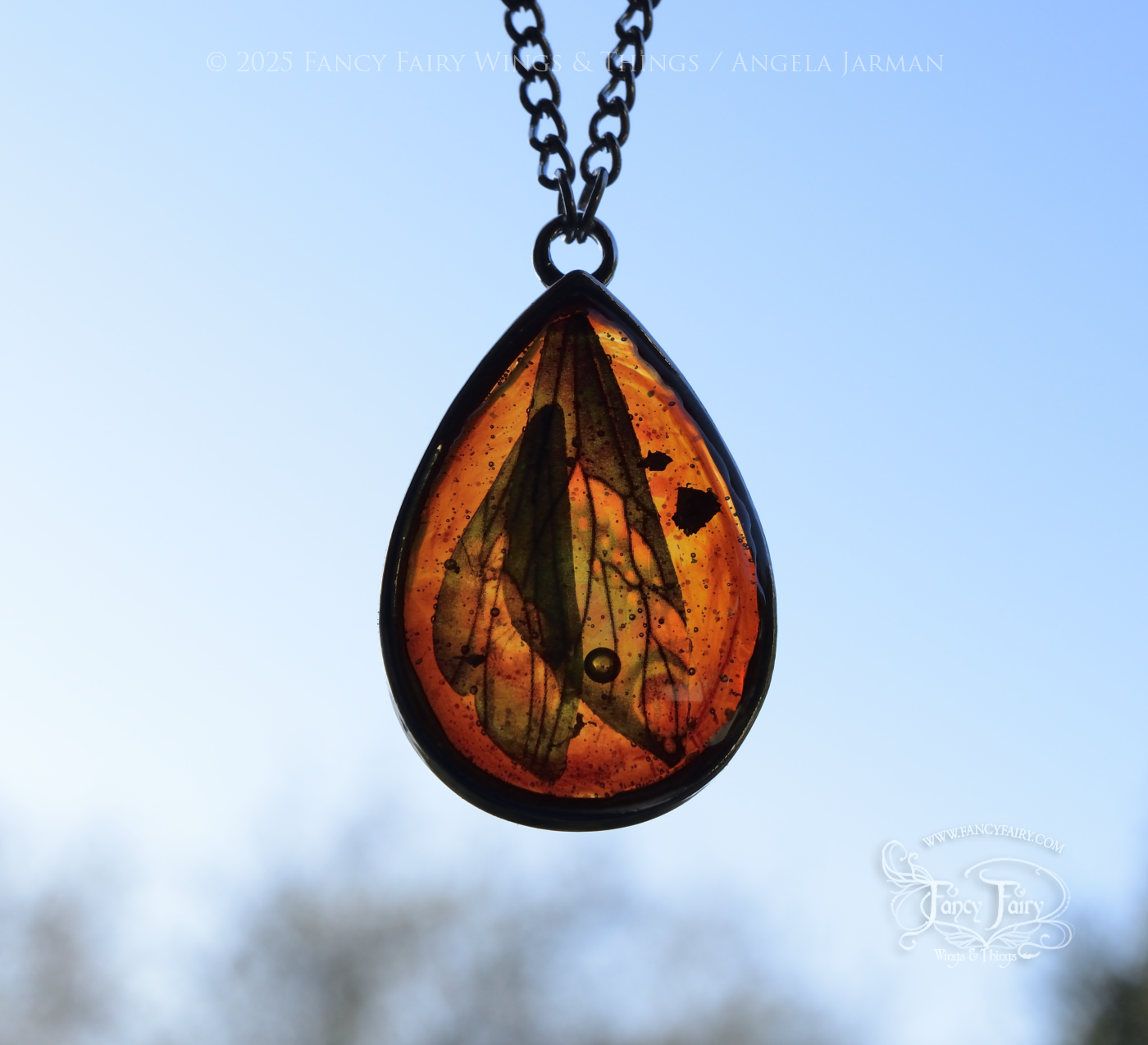 faux amber pendant with teal iridescent fairy wings trapped inside, hanging by silver plated chain, back lit and hanging in front of a bright light blue sky