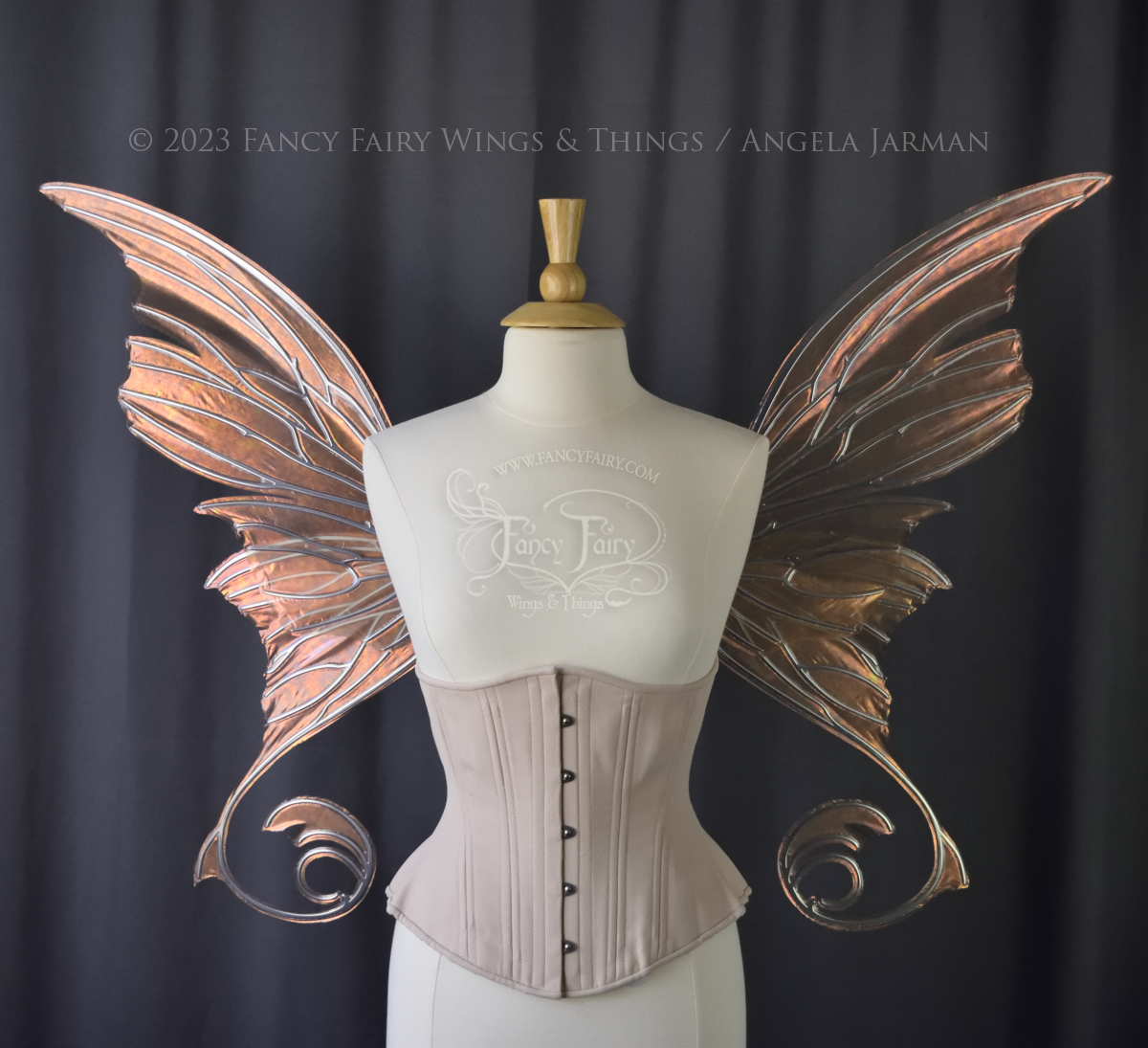 Front view of an ivory dress form wearing an alabaster underbust corset and large rose gold iridescent fairy wings with lots of intricate silver veins, some have 'thorns'. Upper panels curve slightly downwards along top edge with pointy tips, bottom panels have tails. Dark grey background