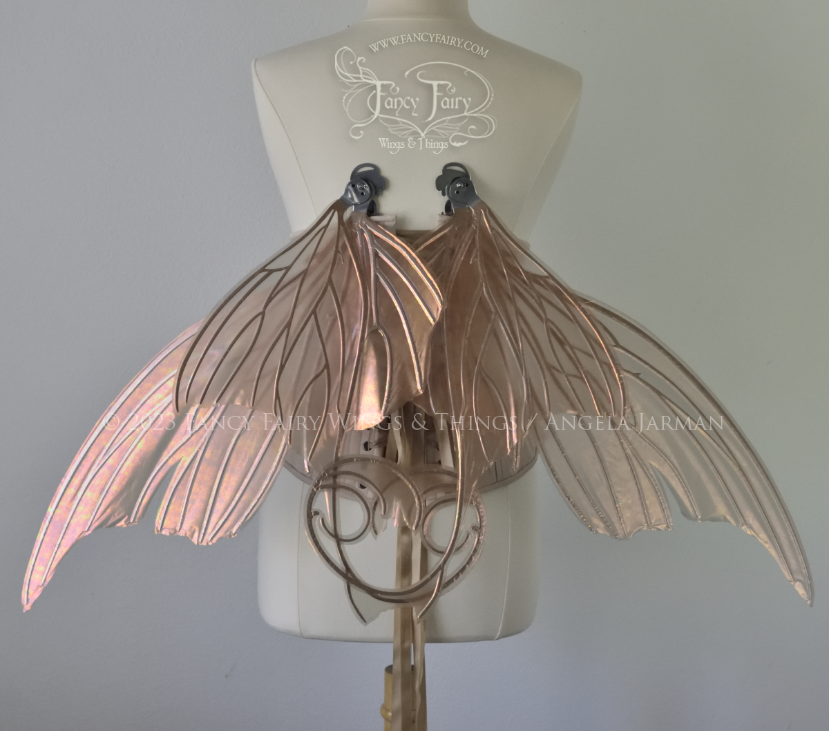 Back view of an ivory dress form wearing an alabaster underbust corset and large rose gold iridescent fairy wings with lots of intricate silver veins, some have 'thorns'. Upper panels curve slightly downwards along top edge with pointy tips, bottom panels have tails, in resting position