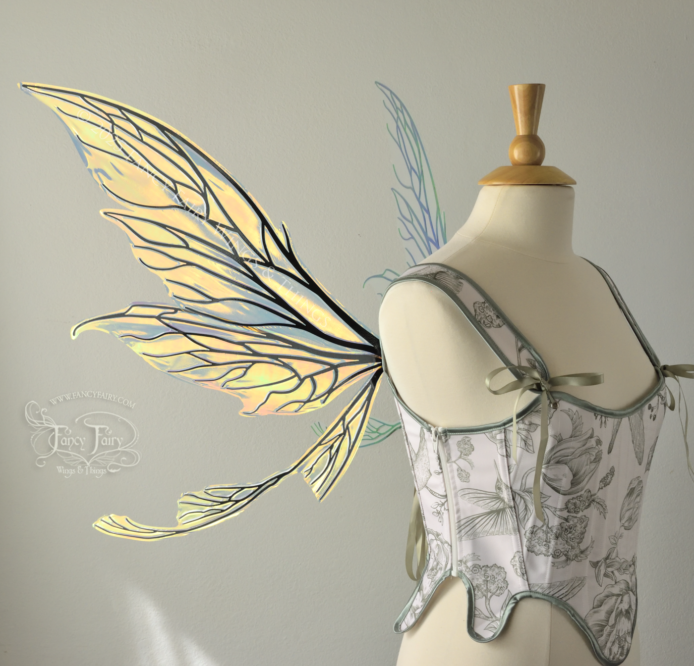 Transparent & iridescent fairy wings reflecting yellow, orange & green with black veins, upper and lower panels are elongated with pointed tips, & curved up ‘tail’ on bottom, worn on a dress form, left side view