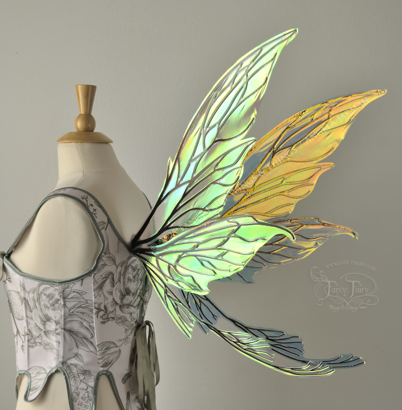 Transparent & iridescent fairy wings reflecting yellow, orange & green with black veins, upper and lower panels are elongated with pointed tips, & curved up ‘tail’ on bottom, worn on a dress form, back 3/4 view