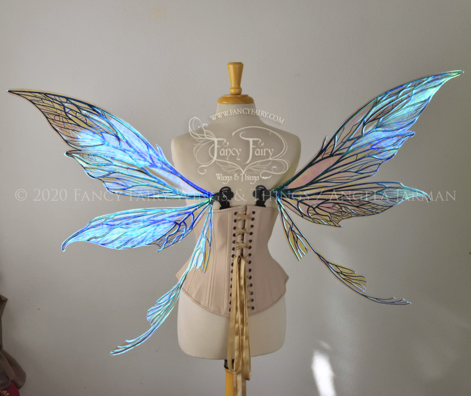Back view of an ivory dress form wearing an alabaster underbust corset and large blue opal iridescent fairy wings with black veins. Upper panels are elongated with pointed tips, curved ‘tail’, lots of thin vein detail