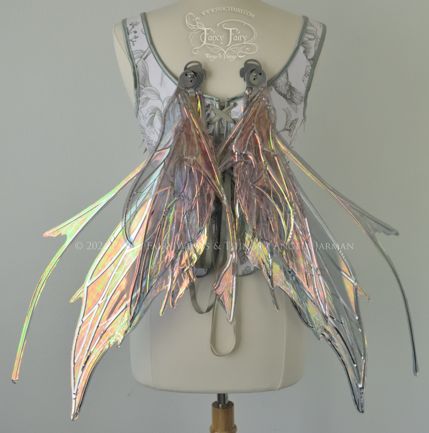 Back view of a dress form wearing a white & green corset and large oil slick colored iridescent fairy wings with antennae on top, 3 panels each side, upper panels come to a point, bottom panels have tails, spikey, detailed silver veins, in resting position