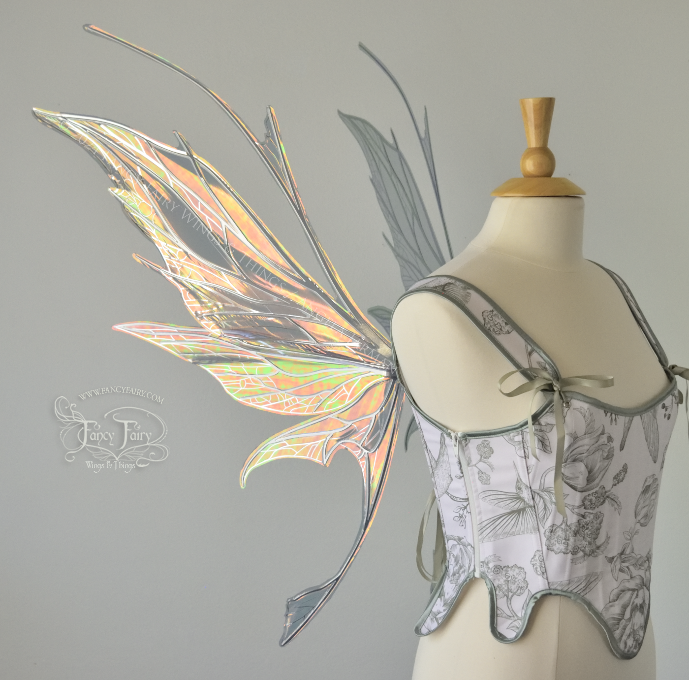 Left side view of a dress form wearing a white & green corset and large oil slick colored iridescent fairy wings with antennae on top, 3 panels each side, upper panels come to a point, bottom panels have tails, spikey, detailed silver veins