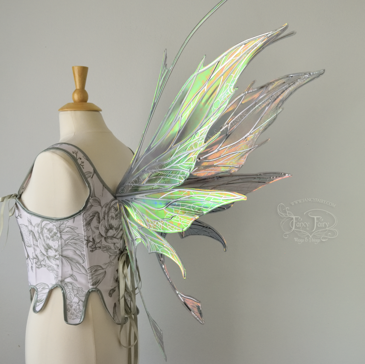 Back 3/4 view of a dress form wearing a white & green corset and large oil slick colored iridescent fairy wings with antennae on top, 3 panels each side, upper panels come to a point, bottom panels have tails, spikey, detailed silver veins