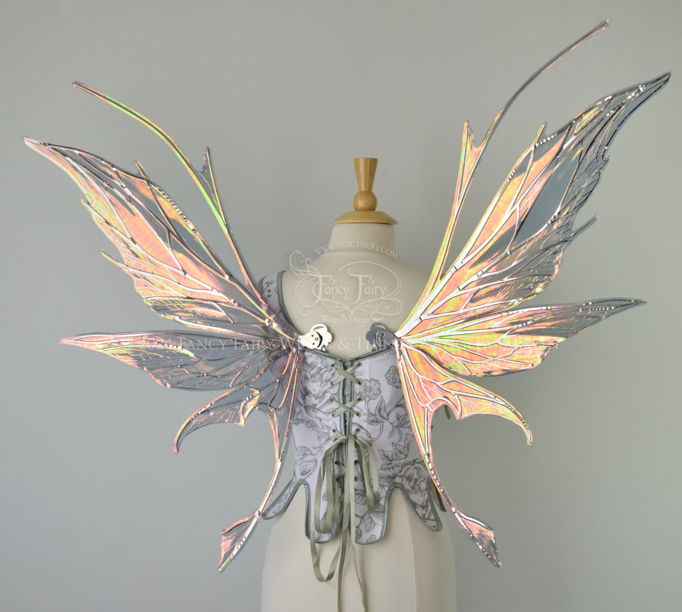 Back view of a dress form wearing a white & green corset and large oil slick colored iridescent fairy wings with antennae on top, 3 panels each side, upper panels come to a point, bottom panels have tails, spikey, detailed silver veins