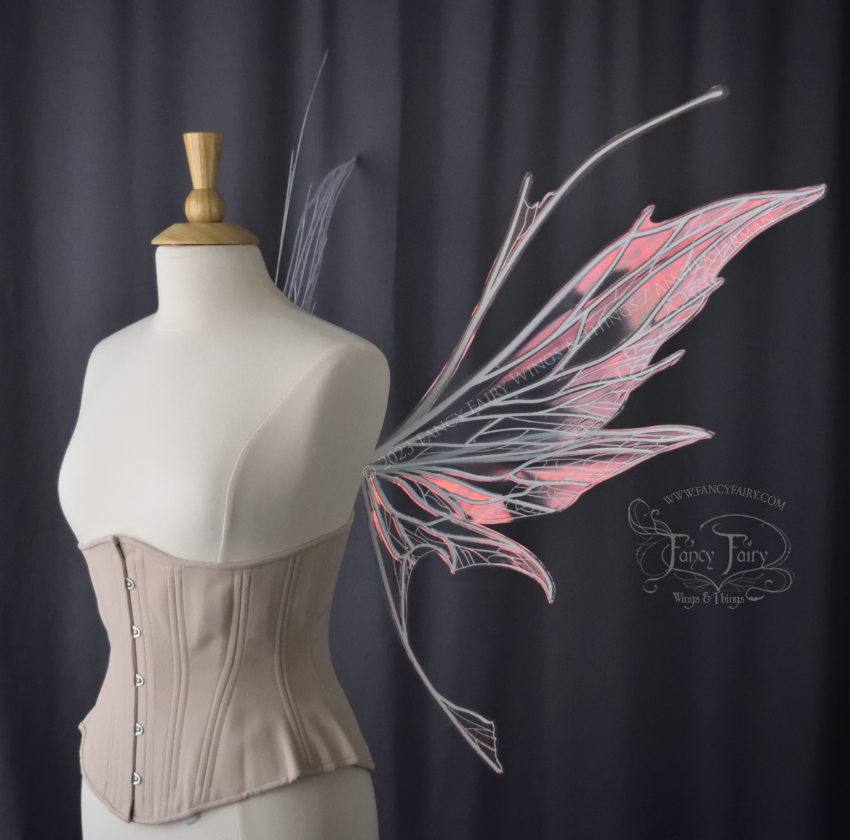 Right side view of an ivory dress form wearing an alabaster underbust corset and large pink iridescent fairy wings with antennae on top. Upper panels come to a point, bottom panels have tails. Spikey, detailed white veins, plain dark grey background