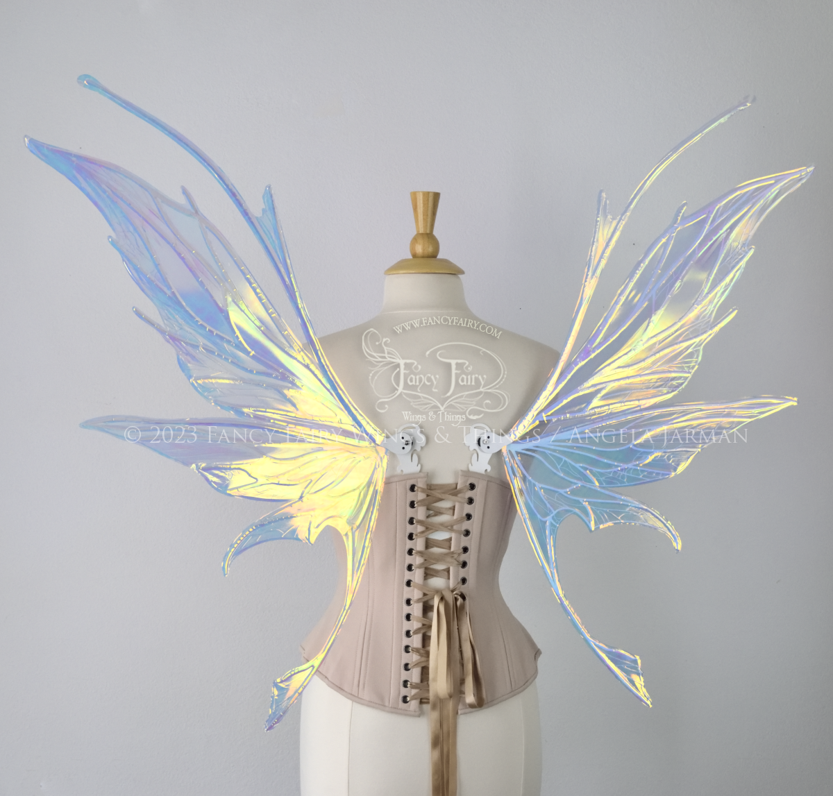 Back view of an ivory dress form wearing an alabaster underbust corset and large multicolor iridescent fairy wings featuring antennae along the top. Upper panels come to a point as do the middle panels, bottom panels have tails. Spikey, detailed white veins. The background is white / light grey