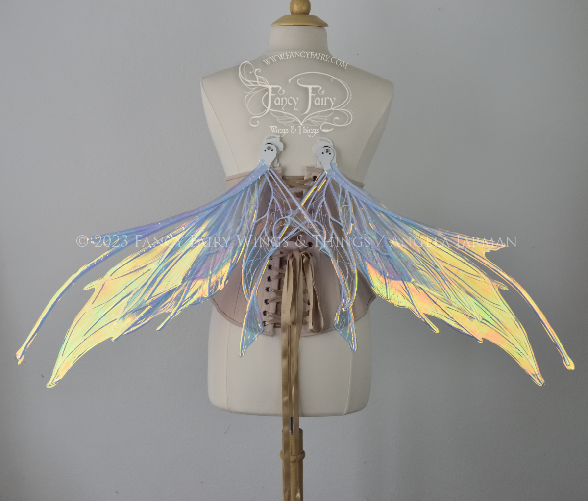 Back view of an ivory dress form wearing an alabaster underbust corset and large multicolor iridescent fairy wings in resting position, featuring antennae along the top. Upper panels come to a point as do the middle panels, bottom panels have tails. Spikey, detailed white veins. The background is white / light grey