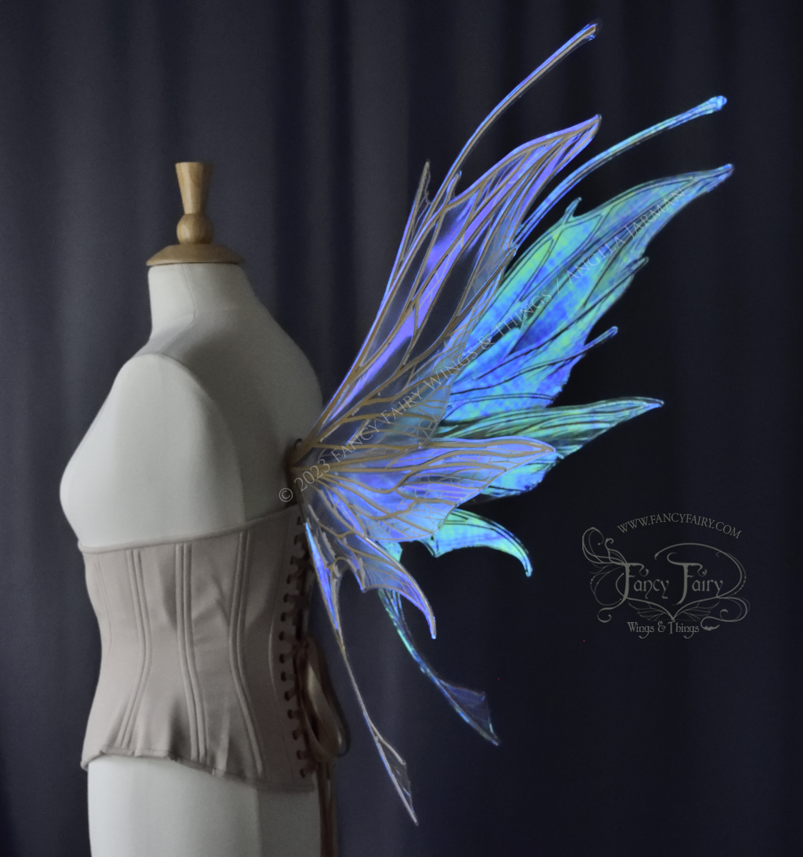 Back 3/4 view of a dress form wearing an underbust corset & large blue/green iridescent fairy wings with antennae, gold veins, spikey shapes, grey background