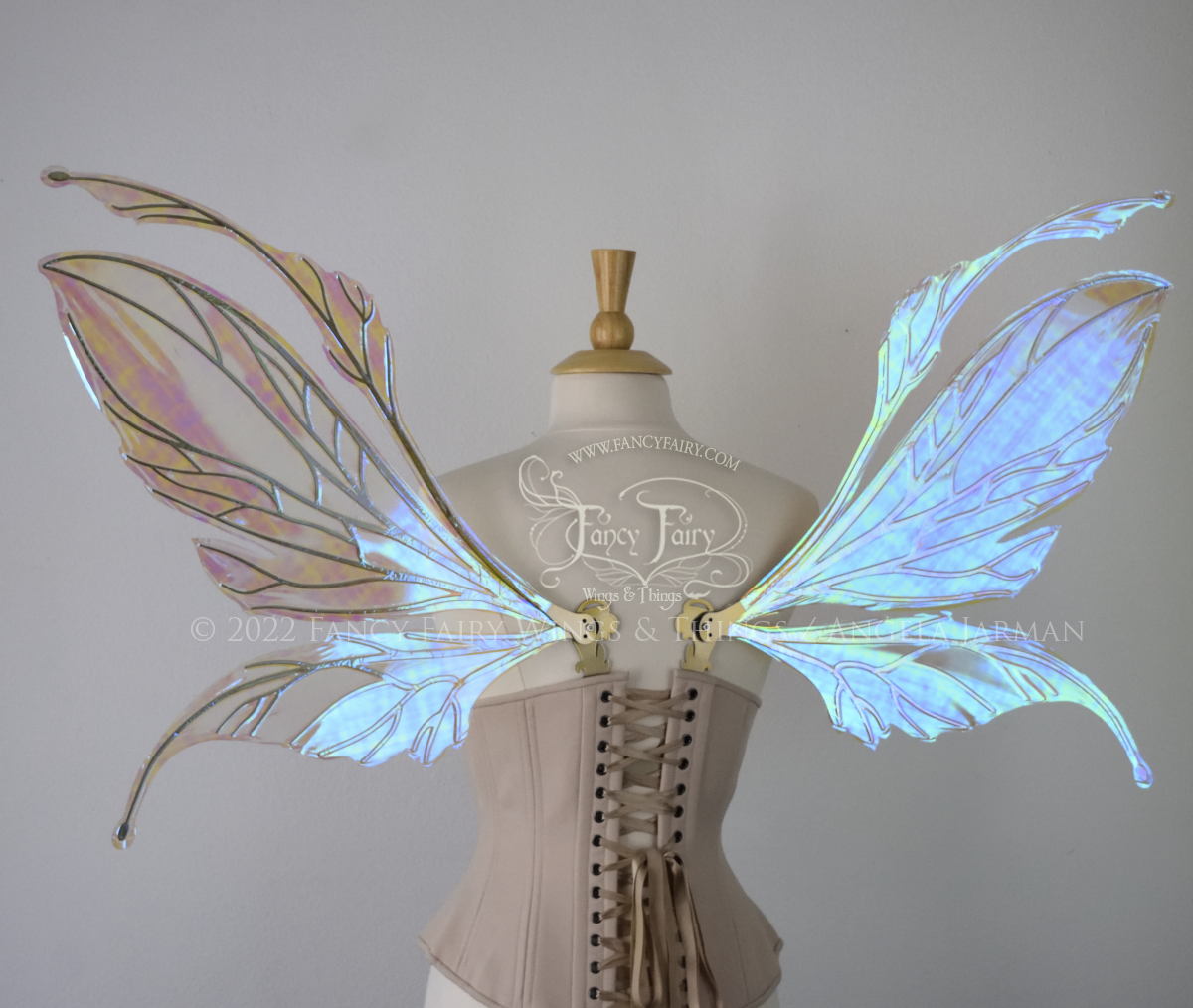 Back view of an ivory dress form wearing an alabaster underbust corset and large fairy wings featuring antennae along the top. The upper panels are elongated with semi-pointed tips, the lower panels are smaller with slender tips that curve downward. They are transparent iridescent with a mostly blue shine with some green and the veins are metallic gold. The background is plain white and my logo and copyright notice are visible.