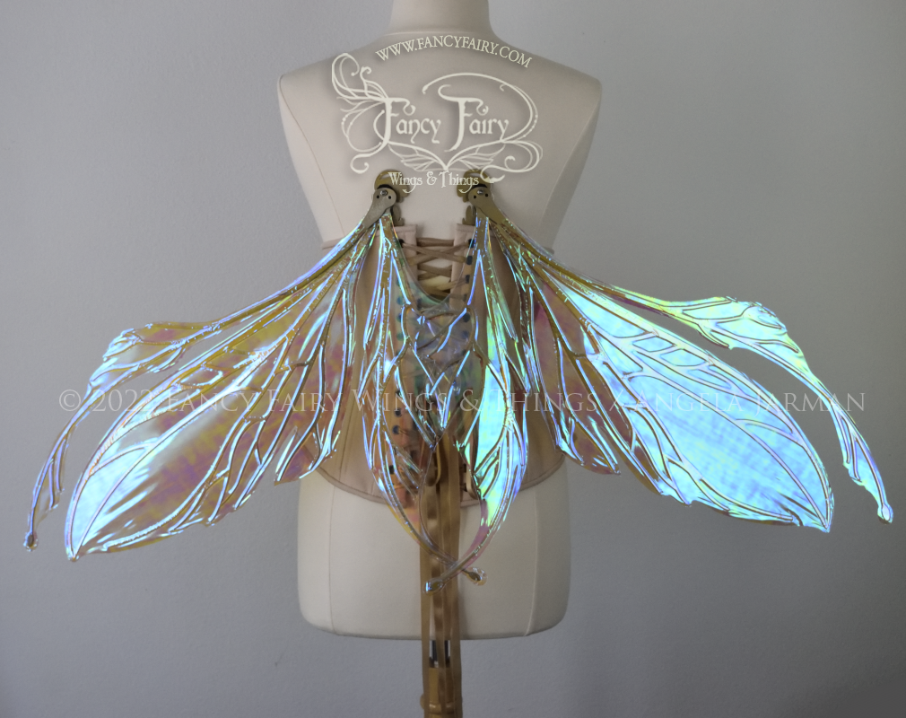 Back view of an ivory dress form wearing an alabaster underbust corset and large fairy wings (in resting position) featuring antennae along the top. The upper panels are elongated with semi-pointed tips, the lower panels are smaller with slender tips that curve downward. They are transparent iridescent with a mostly blue shine with some green and the veins are metallic gold. The background is plain white and my logo and copyright notice are visible.