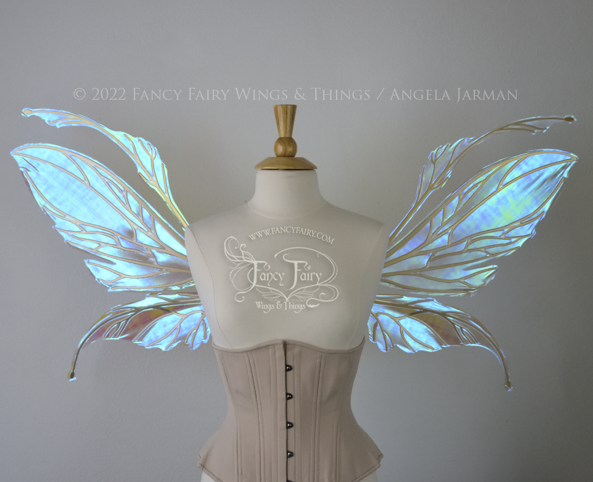 Front view of an ivory dress form wearing an alabaster underbust corset and large fairy wings featuring antennae along the top. The upper panels are elongated with semi-pointed tips, the lower panels are smaller with slender tips that curve downward. They are transparent iridescent with a mostly blue shine with some green and the veins are metallic gold. The background is plain white and my logo and copyright notice are visible.