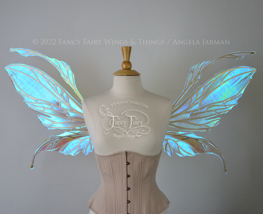 Front view of an ivory dress form wearing an alabaster underbust corset and large fairy wings featuring antennae along the top. The upper panels are elongated with semi-pointed tips, the lower panels are smaller with slender tips that curve downward. They are transparent iridescent with a mostly blue shine with some green and the veins are metallic gold. The background is plain white and my logo and copyright notice are visible.