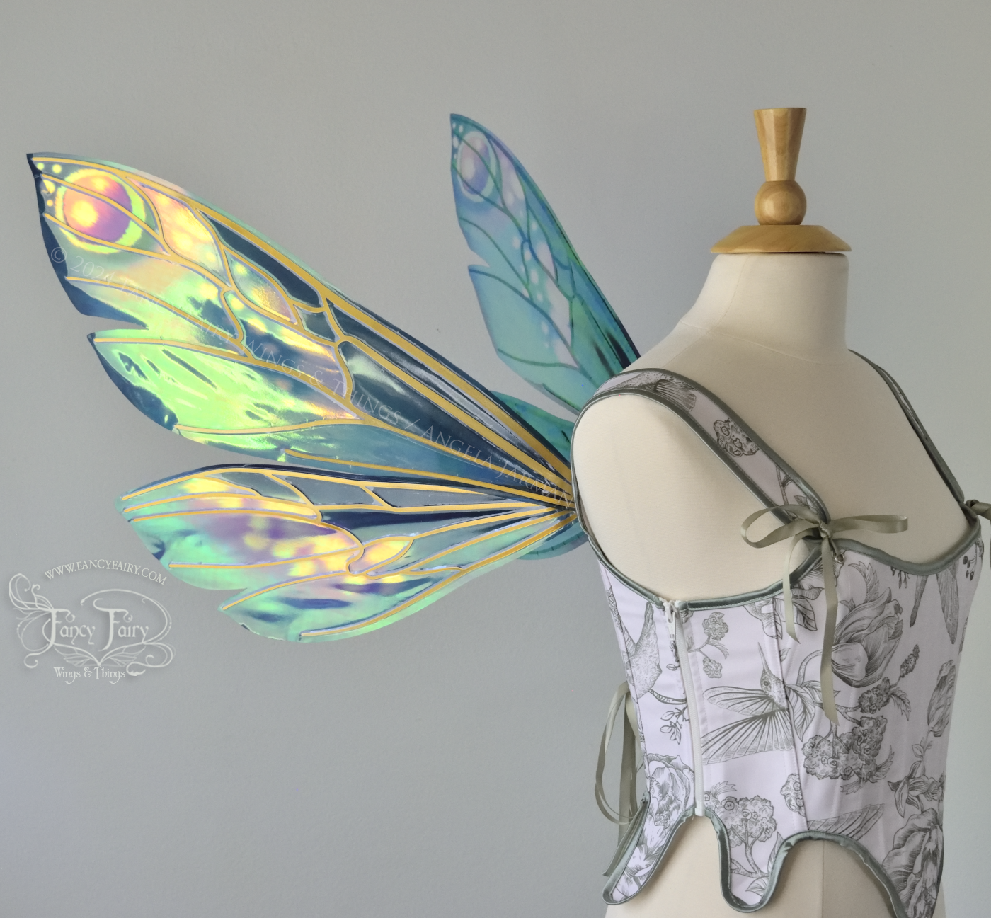 Left side view of iridescent fairy wings in various shades of green, teal, blue, lavender with touches of pink, with gold veins, worn on a dress form with white & green corset