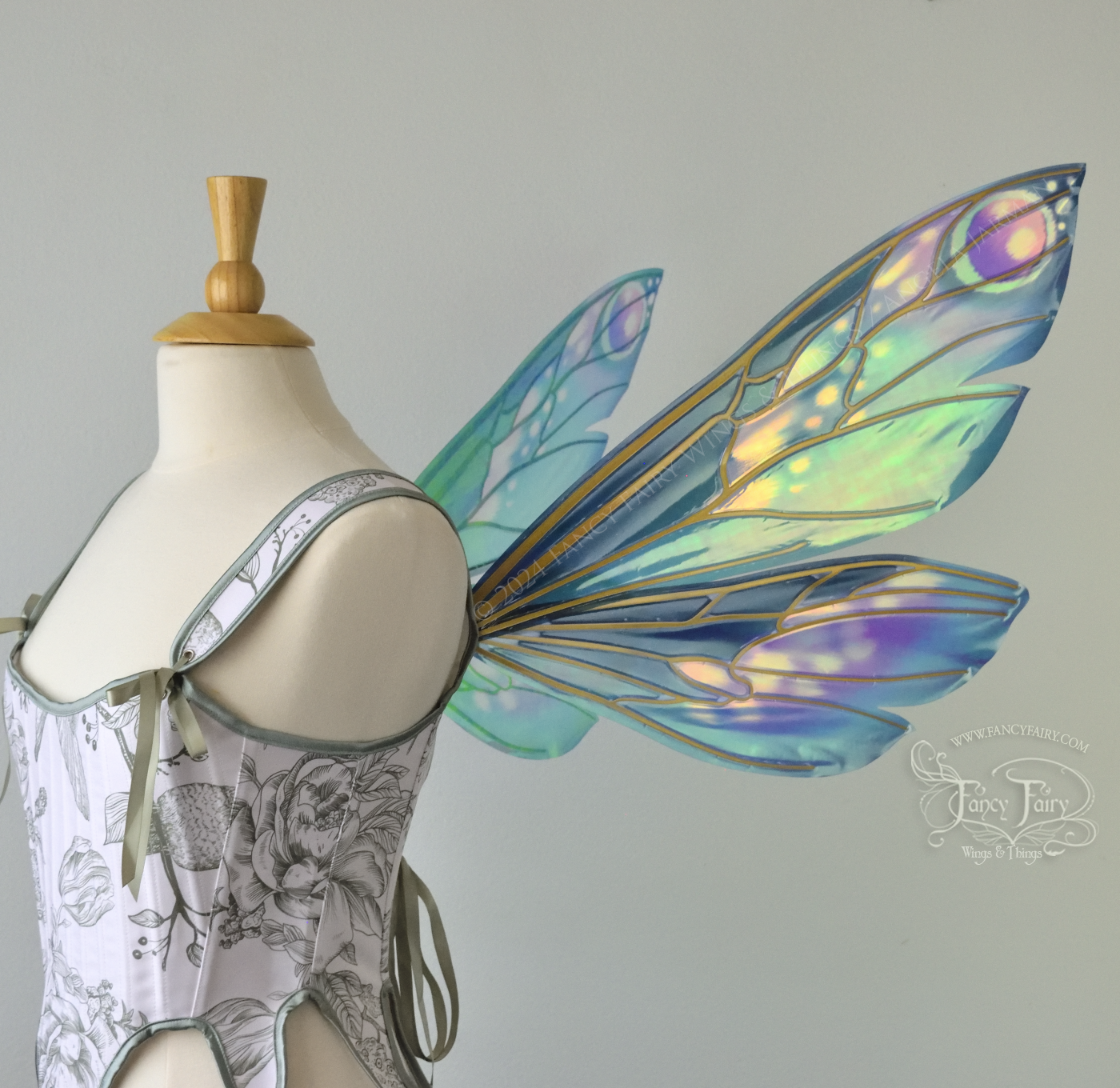 Right side view of iridescent fairy wings in various shades of green, teal, blue, lavender with touches of pink, with gold veins, worn on a dress form with white & green corset