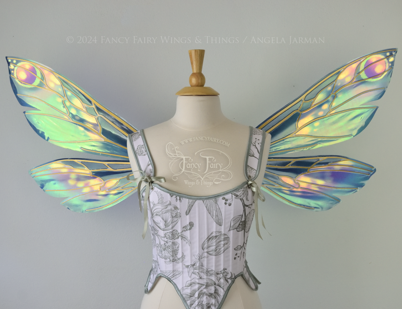 Front view of iridescent fairy wings in various shades of green, teal, blue, lavender with touches of pink, with gold veins, worn on a dress form with white & green corset