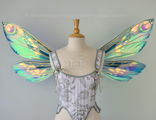 Front view of iridescent fairy wings in various shades of green, teal, blue, lavender with touches of pink, with gold veins, worn on a dress form with white & green corset