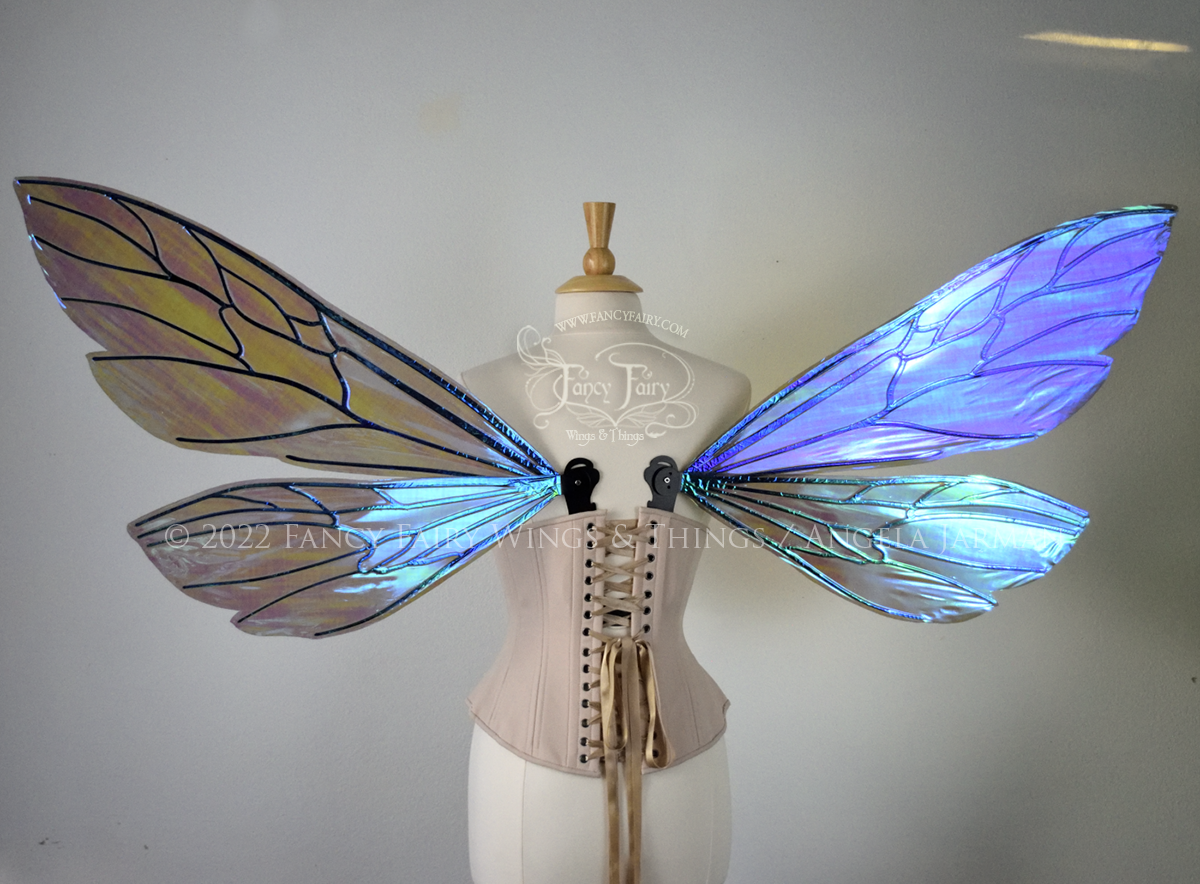 Back view of an ivory dress form wearing an alabaster underbust corset and large fairy wings similar to wasp wings, upper and lower panels are both elongated with rounded and slightly pointed tips. They are iridescent green / blue with some purplish pink undertones in the shadows, with black veins. The background is plain white and my logo and copyright notice are visible.