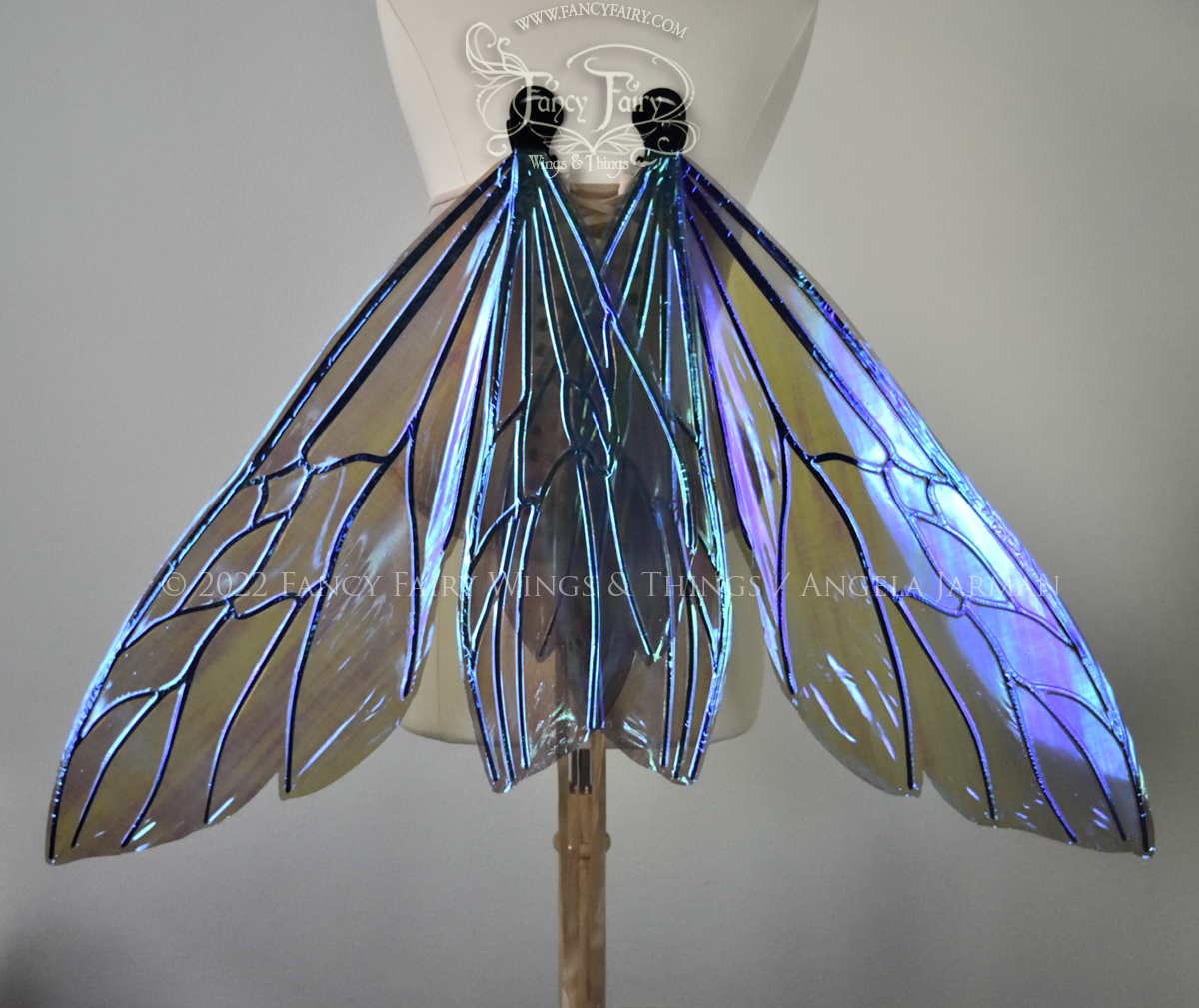 Back view of an ivory dress form wearing an alabaster underbust corset and large fairy wings similar to wasp wings in resting position, upper and lower panels are both elongated with rounded and slightly pointed tips. They are iridescent green / blue with some purplish pink undertones in the shadows, with black veins. The background is plain white and my logo and copyright notice are visible.