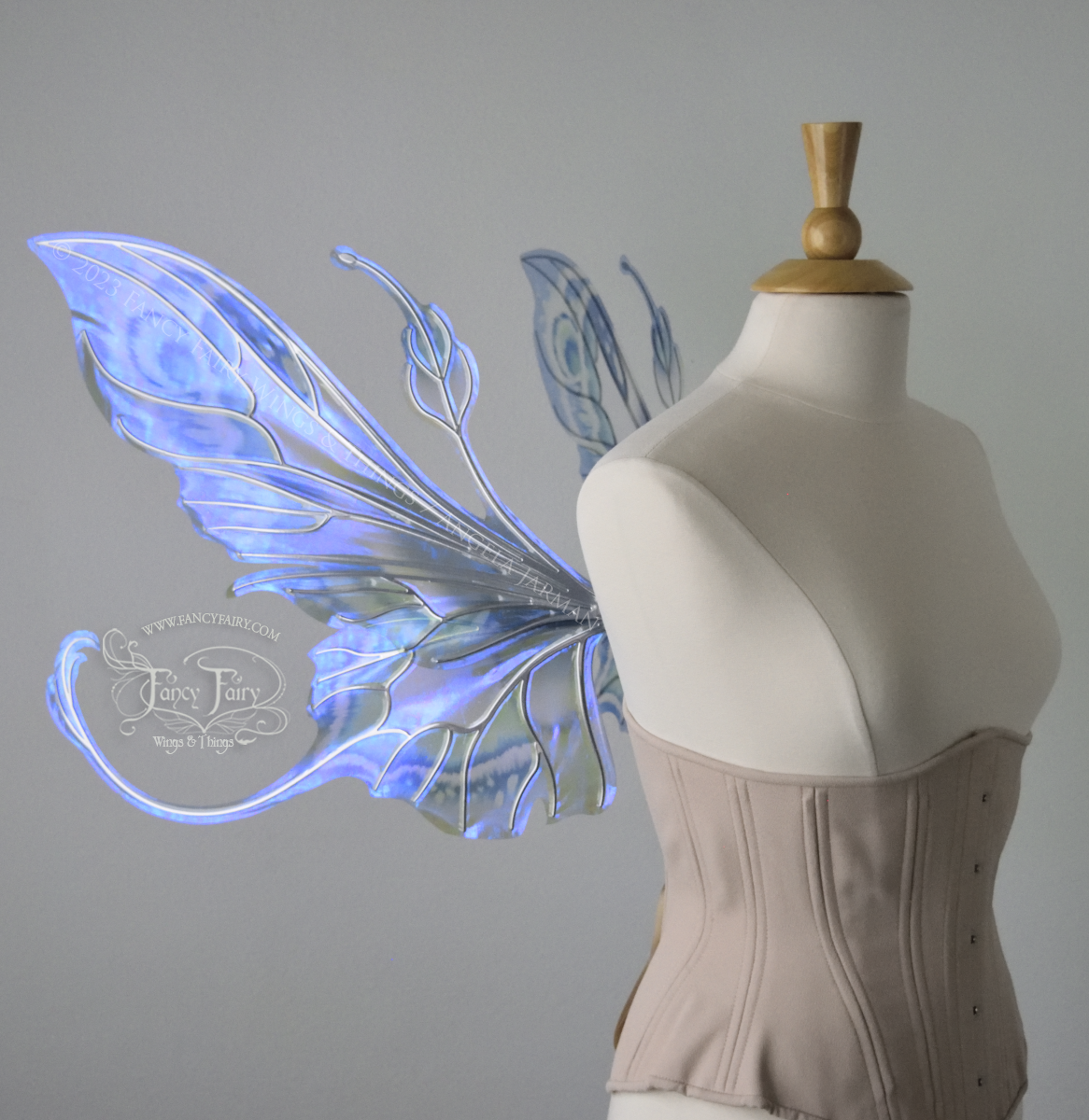 Left side view of large blue, violet & teal painted iridescent fairy wings, elongated upper panels with antennae, bottom panels have a tail curving upwards, silver veins, worn on a dress form