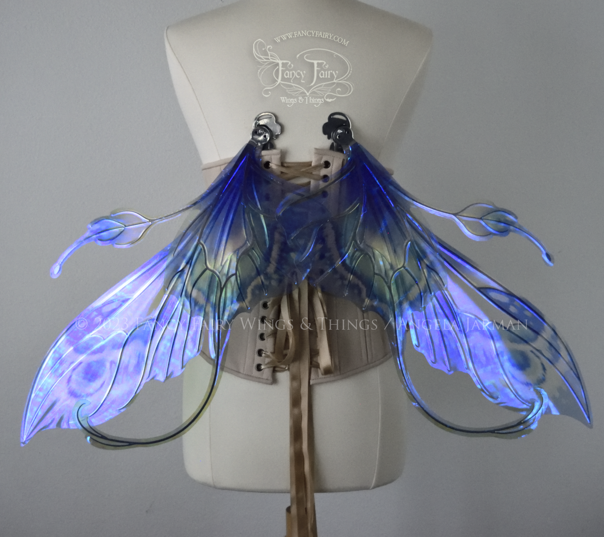 Back view of large blue, violet & teal painted iridescent fairy wings, elongated upper panels with antennae, bottom panels have a tail curving upwards, silver veins, worn on a dress form, in resting position