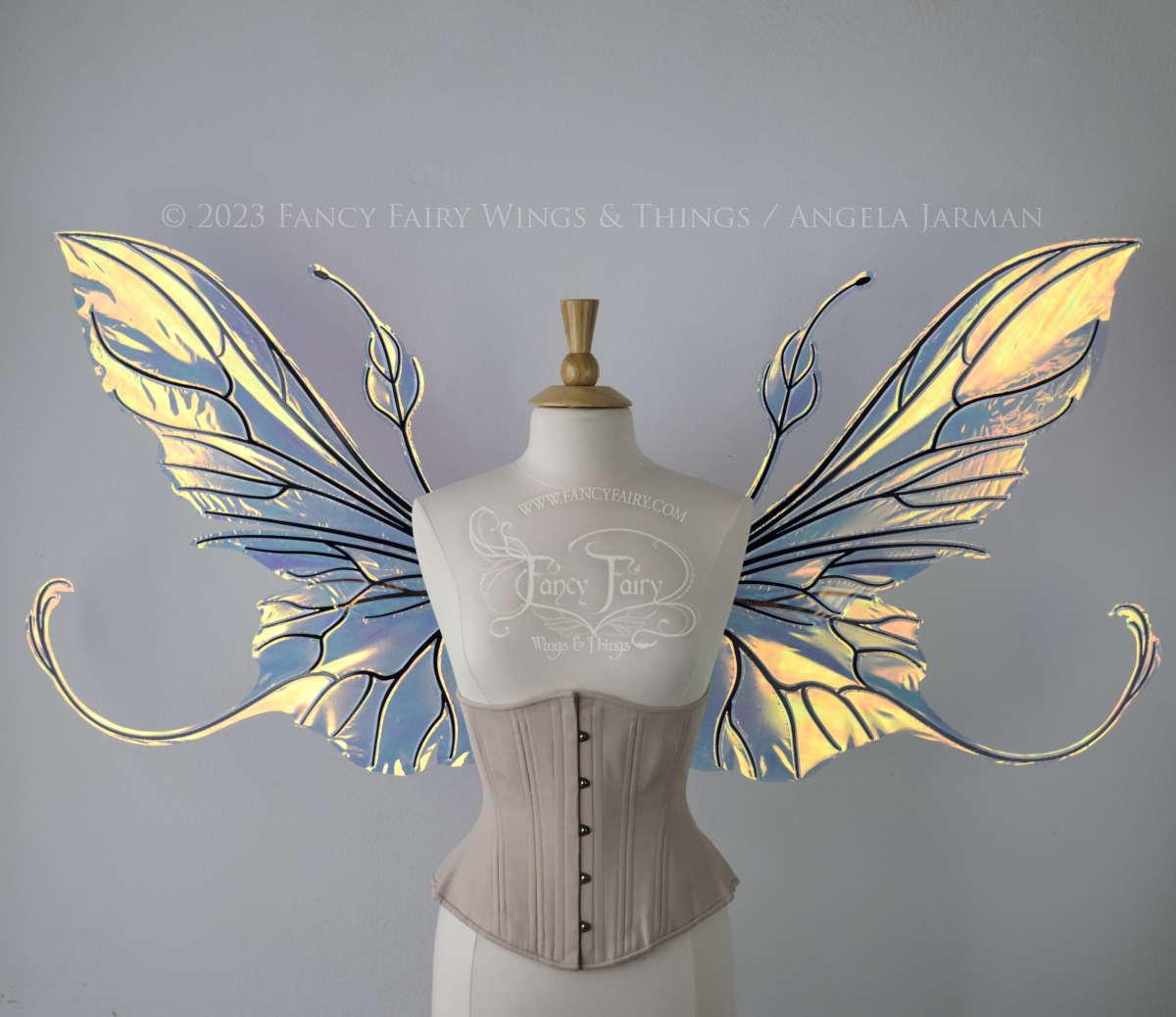 Front view of an ivory dress form wearing an alabaster underbust corset & extra large iridescent fairy wings with elongated upper panels & antennae with bottom panels that have a tail curving upwards, black veins. Background is white / light grey