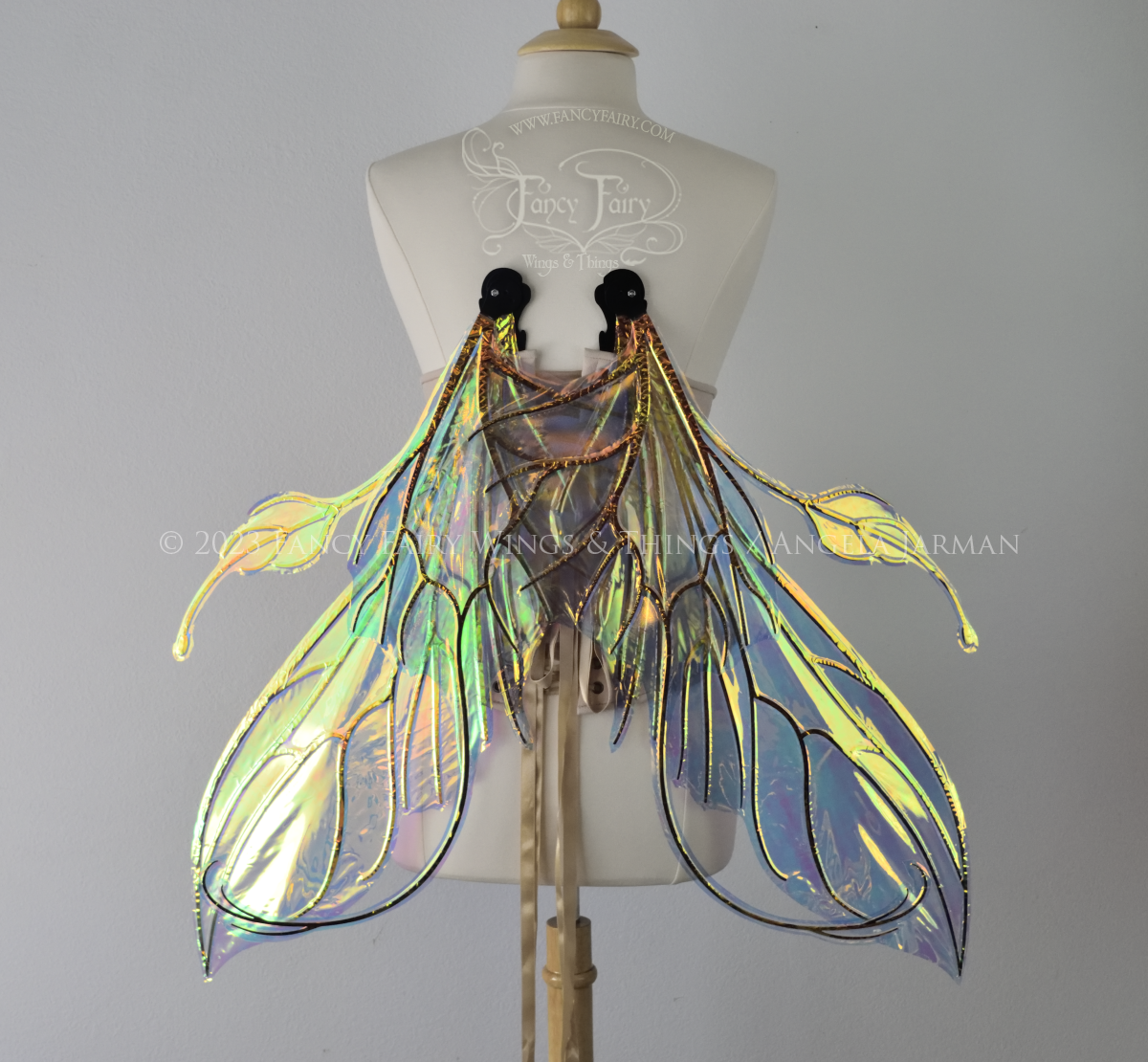 Back view of an ivory dress form wearing an alabaster underbust corset & extra large iridescent fairy wings (in resting position pointed downward) with elongated upper panels & antennae with bottom panels that have a tail curving upwards, black veins. Background is white / light grey