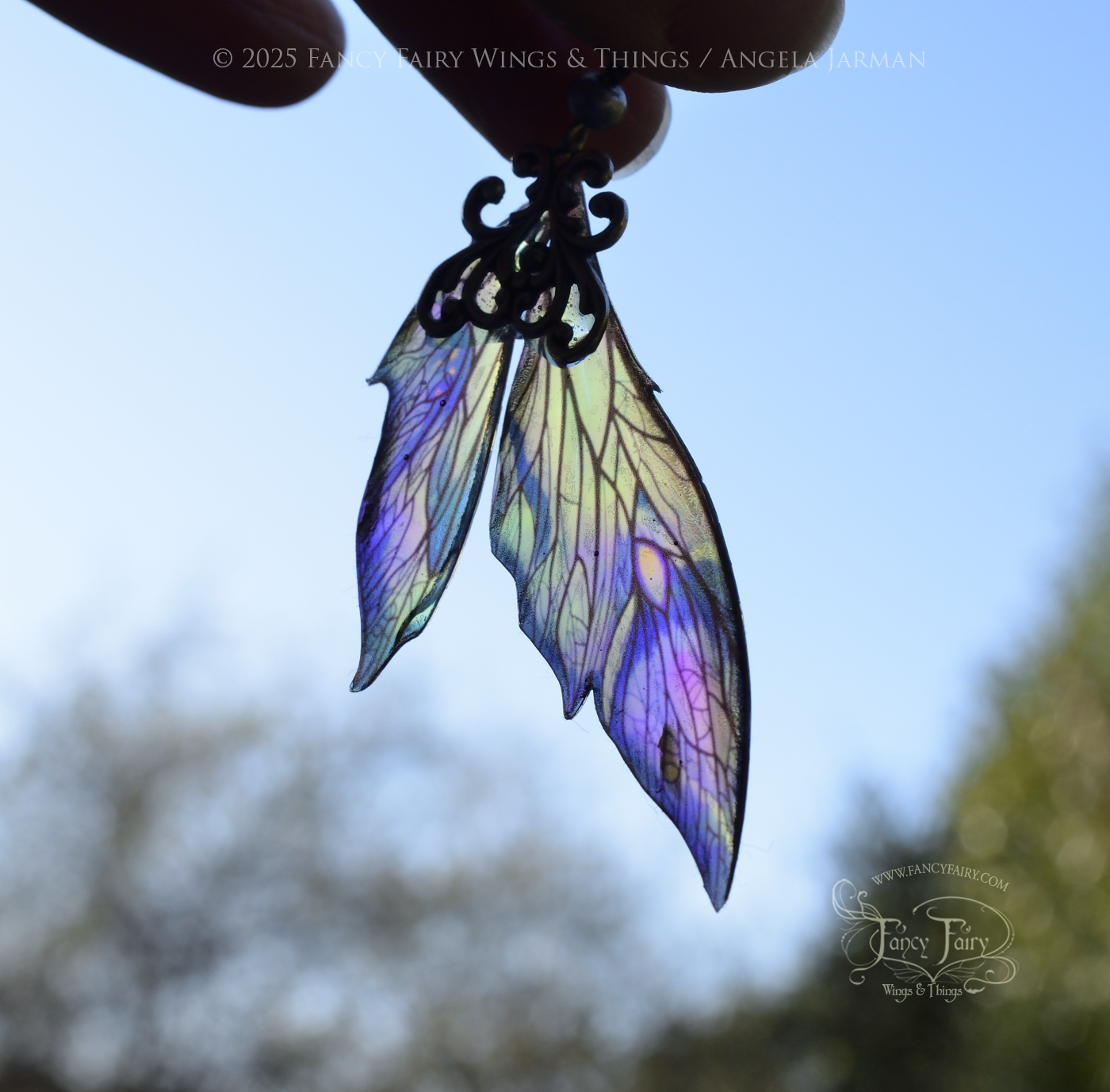 Iridescent fairy wing earrings in blues with silver filigree are back lit and dangle in front of a bright blue sky